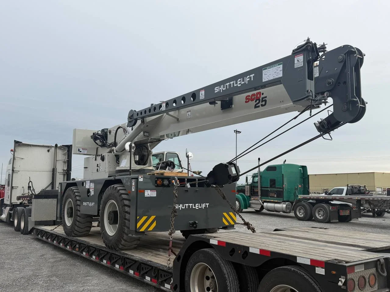 A large industrial shuttle lift crane on a flatbed trailer, with other trucks and equipment in the background outdoors.