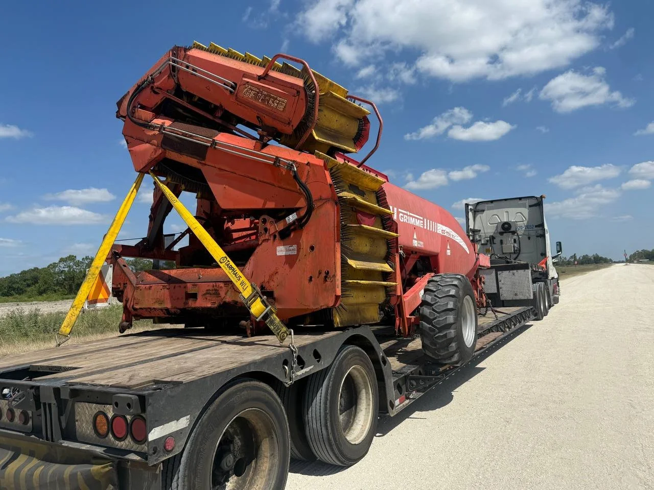 A heavy-duty truck transporting a large piece of industrial equipment on a flatbed trailer on a dirt road under a partly cloudy sky.