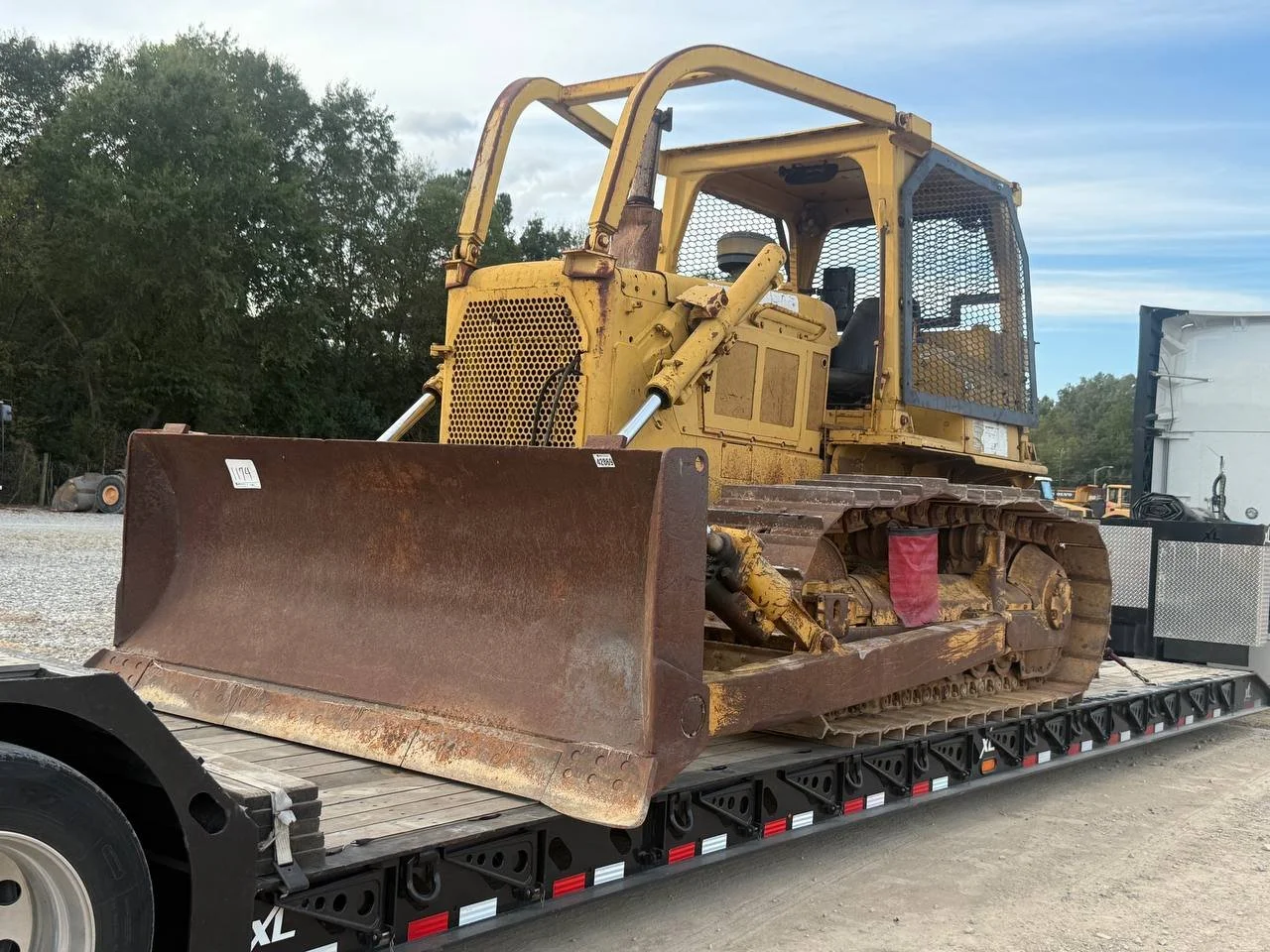 A large yellow bulldozer with a rusted blade is loaded on a flatbed trailer, surrounded by trees and a cloudy sky.