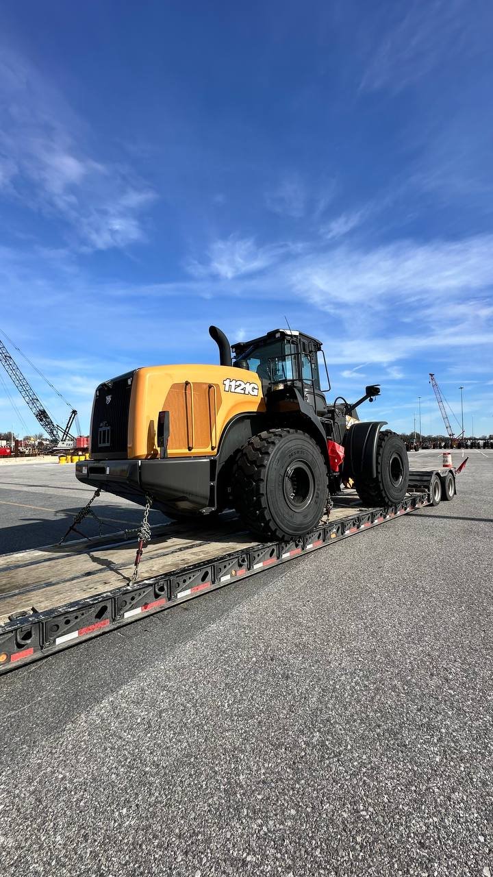 A large yellow and black industrial machine, possibly a bulldozer or loader, secured on a flatbed trailer in an outdoor lot with cranes and construction equipment in the background under a blue sky.