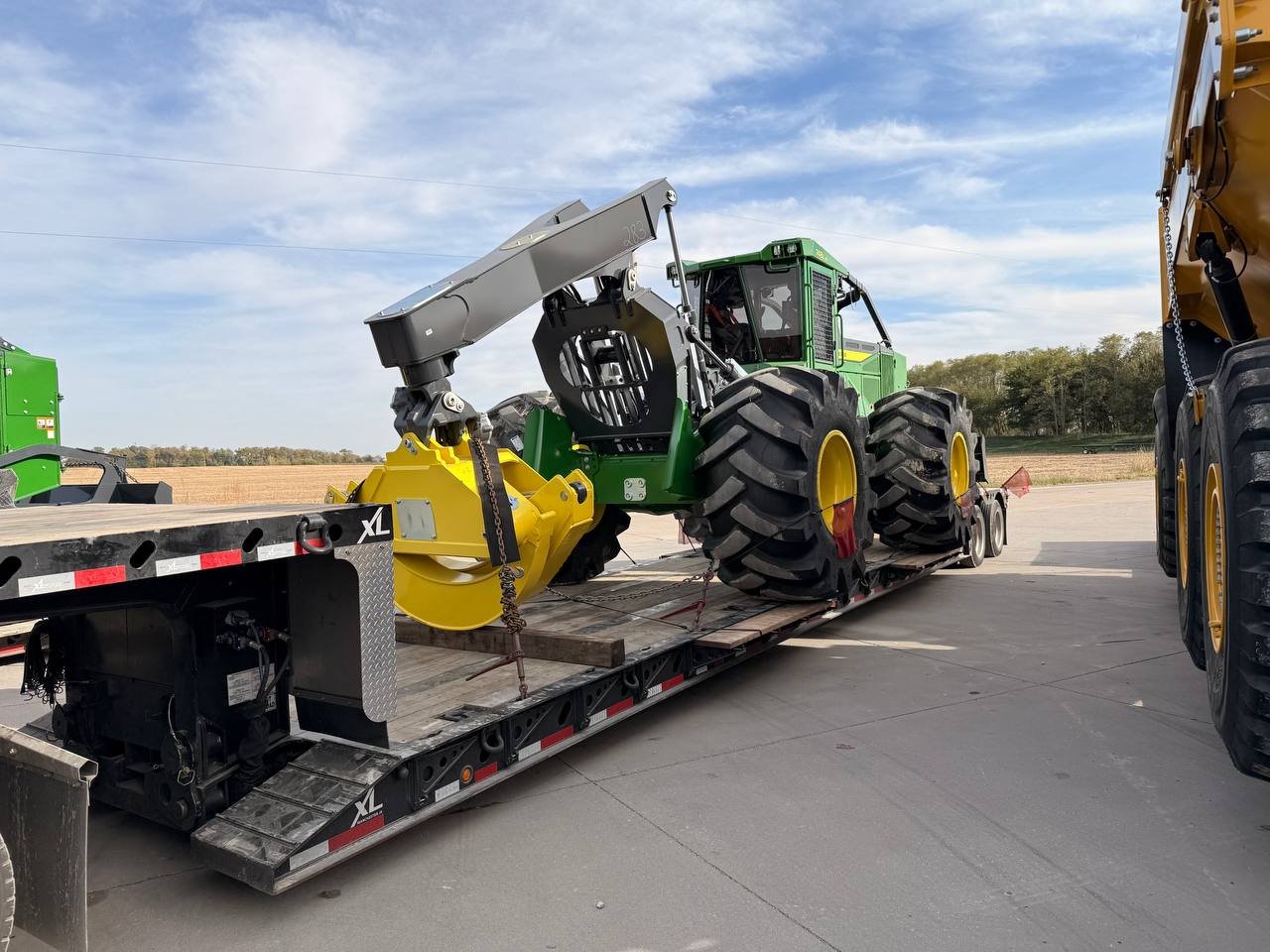 A green John Deere tractor with large tires and yellow rims is loaded on a flatbed trailer, secured with chains, against a rural background with open fields and a partly cloudy sky.