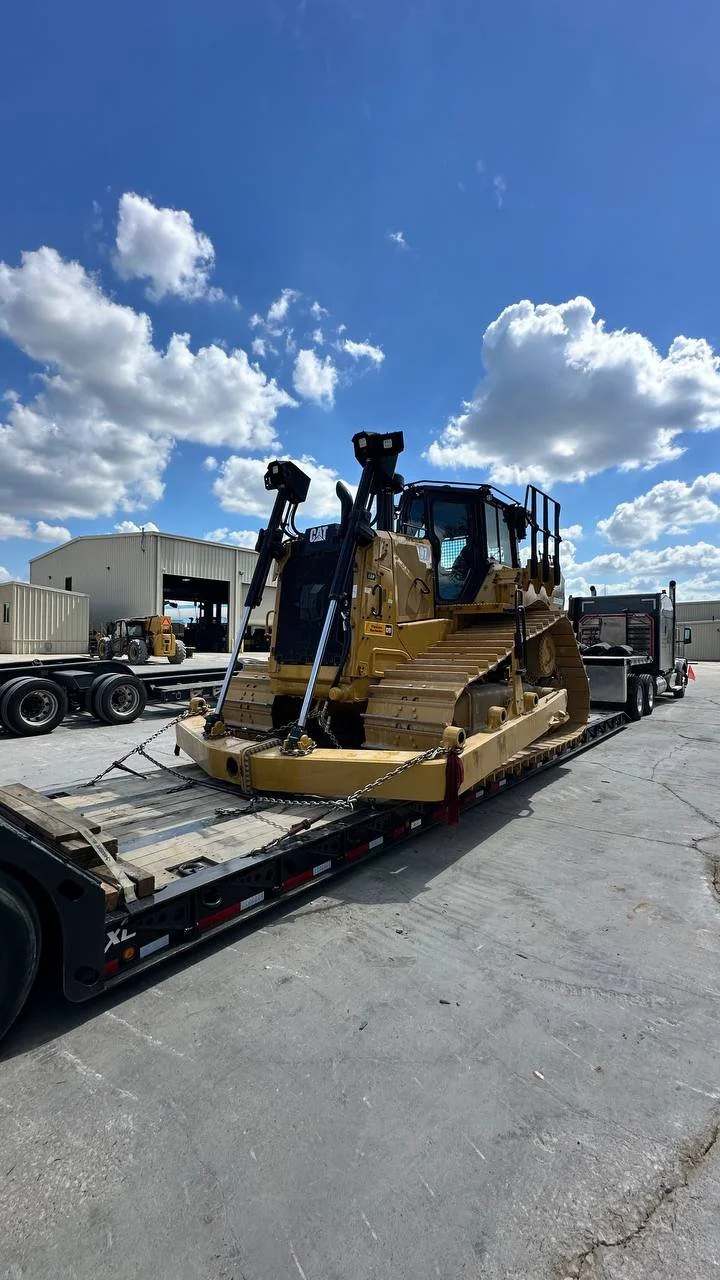 A large yellow bulldozer loaded onto a flatbed trailer in an industrial yard with other construction equipment and storage buildings, under a blue sky with scattered clouds.