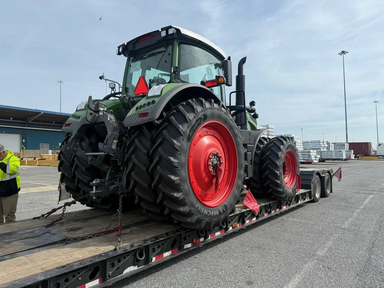 Large green tractor with red wheels loaded on a flatbed truck in an outdoor parking lot with industrial buildings and stacked pallets in the background.