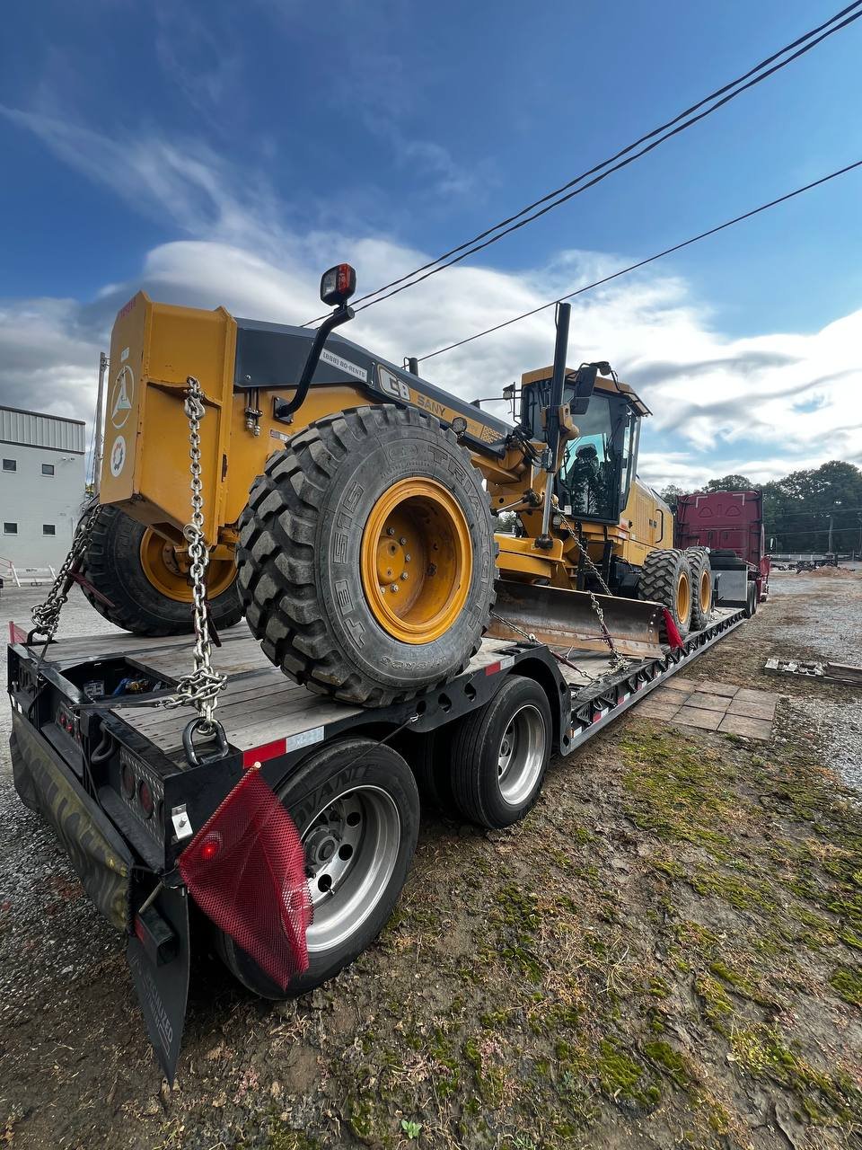 A large yellow construction machine, specifically a road roller, is secured on a flatbed trailer for transportation. The truck is parked outside on a gravel and dirt surface with a cloudy sky overhead.