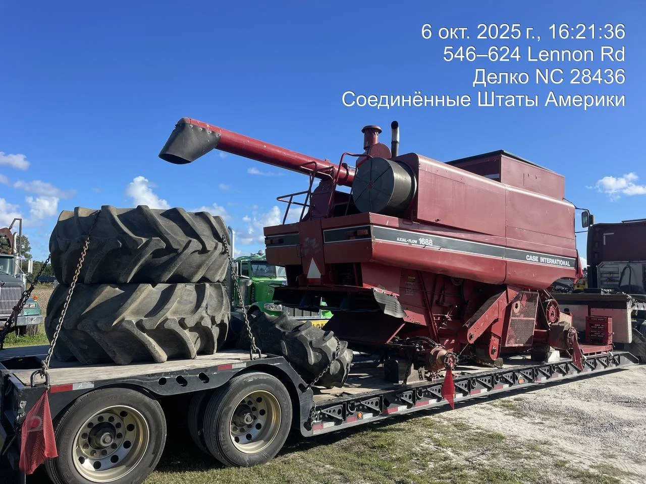 A large red agricultural combine harvester is on a flatbed trailer, with large tractor tires stacked on the trailer. There are other vehicles in the background, and a bright blue sky with some clouds.