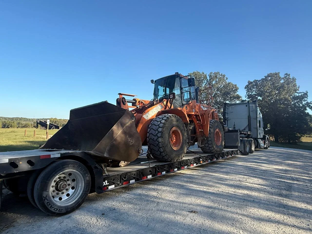 Large orange loader on a flatbed truck, parked outdoors on a gravel surface with trees and a clear blue sky in the background.