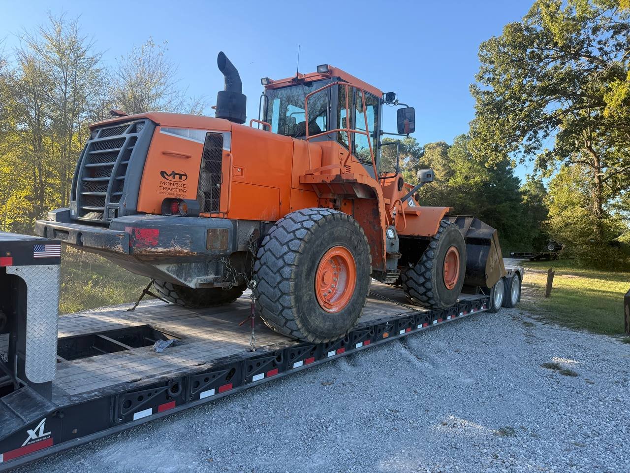 An orange tractor loaded on a flatbed trailer on a gravel road with trees and a clear blue sky in the background.