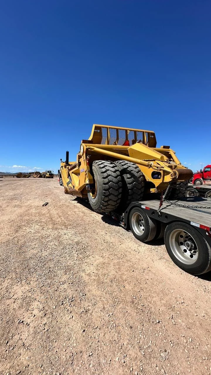 A large yellow heavy machinery piece loaded on a flatbed trailer in an open dirt area, with a blue sky overhead.