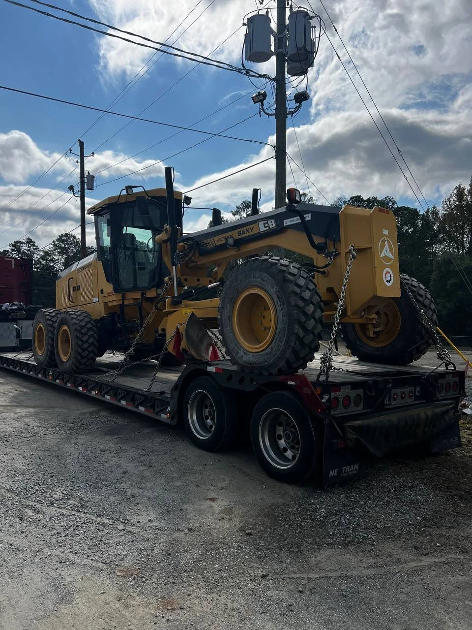 A yellow heavy construction grader machine secured on a flatbed trailer, parked outdoors with a partly cloudy sky and power lines in the background.
