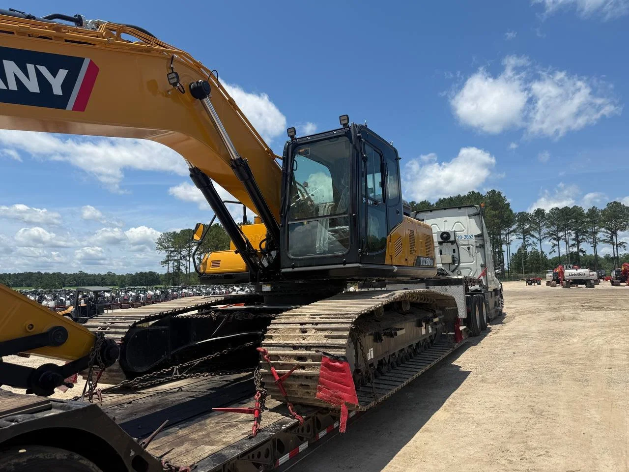 A large yellow and black excavator loaded on a flatbed truck in a construction yard, with a line of golf carts in the background under a blue sky with clouds.