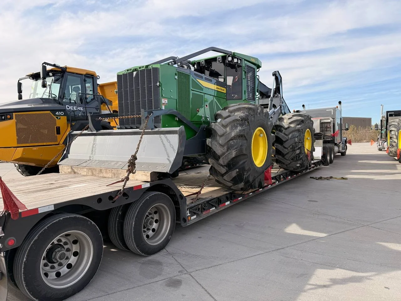 A flatbed trailer carrying a green John Deere tractor with large yellow wheels and a snow plow attachment, with a yellow and black Deere bulldozer visible in the background, on a concrete lot under a partly cloudy sky.