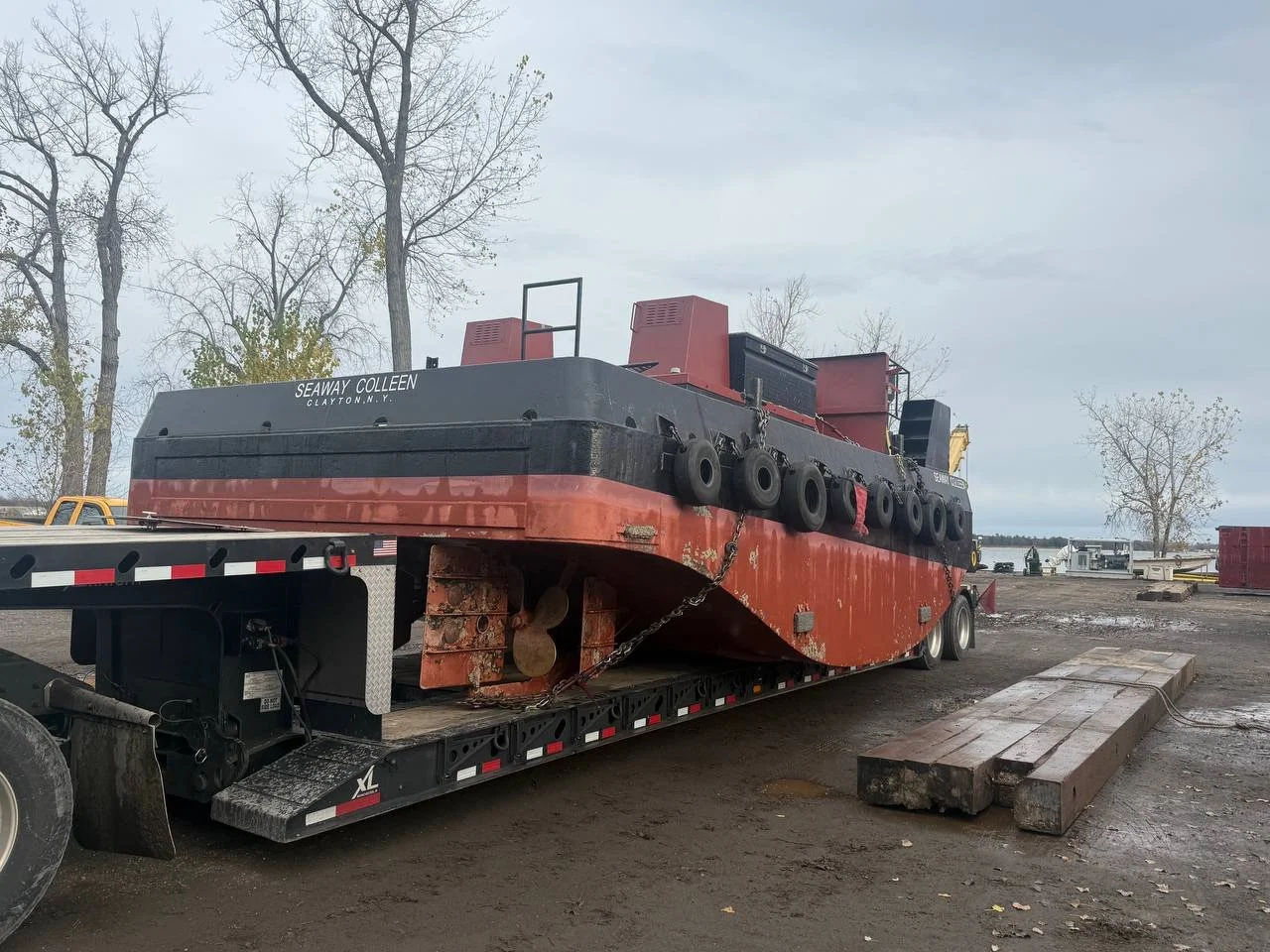 A large tugboat named Seaway Colleen is loaded on a flatbed trailer outdoors. The boat is black and red with multiple tires along its side for bumping. The background shows trees and an overcast sky.