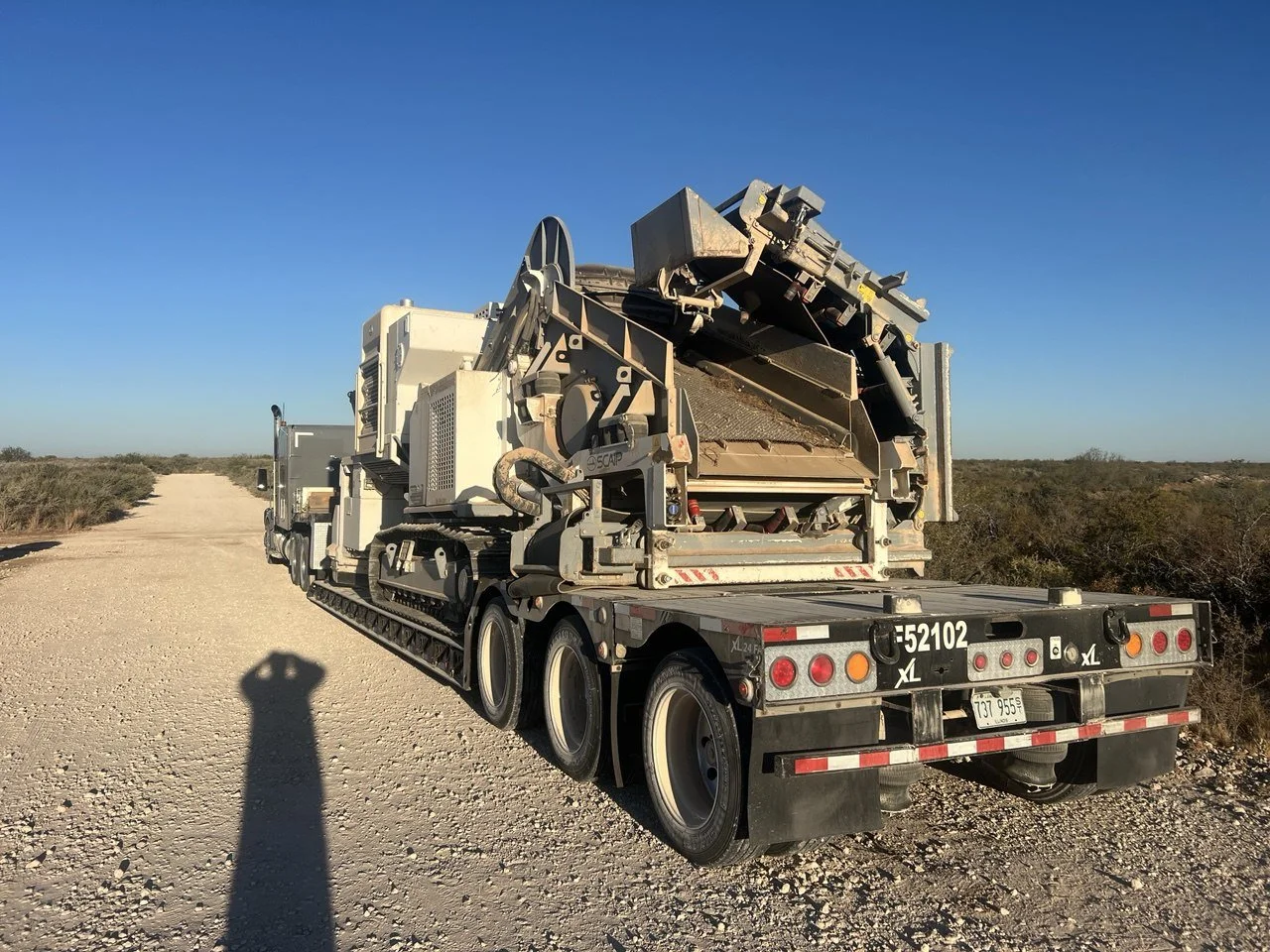 A large flatbed truck carrying industrial machinery is parked on a dirt road in a desert landscape with sparse bushes and a clear blue sky.