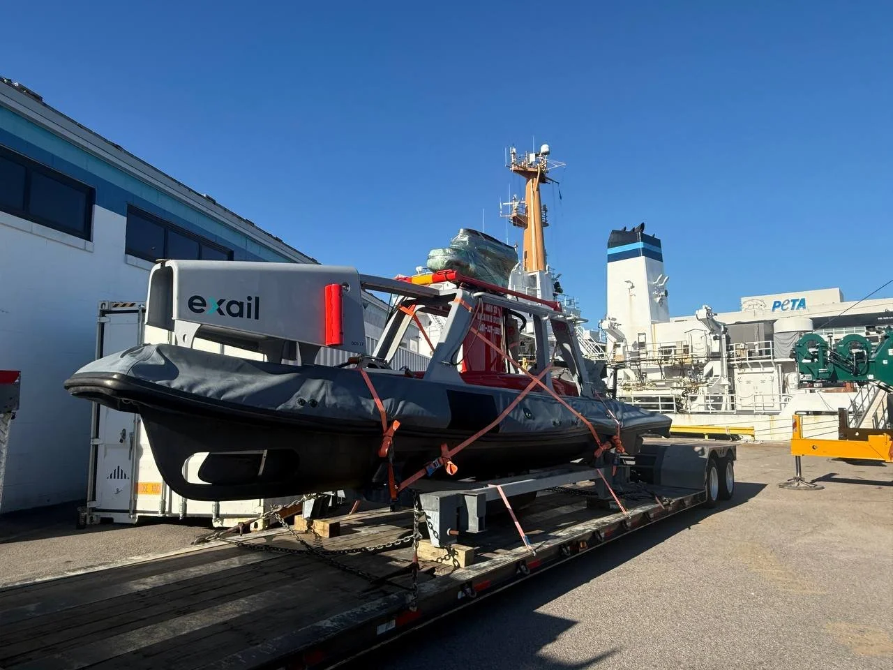 A Black rescue boat loaded on a flatbed trailer, secured with orange straps, outside a large building with a ship and a tall structure in the background, on a clear day.