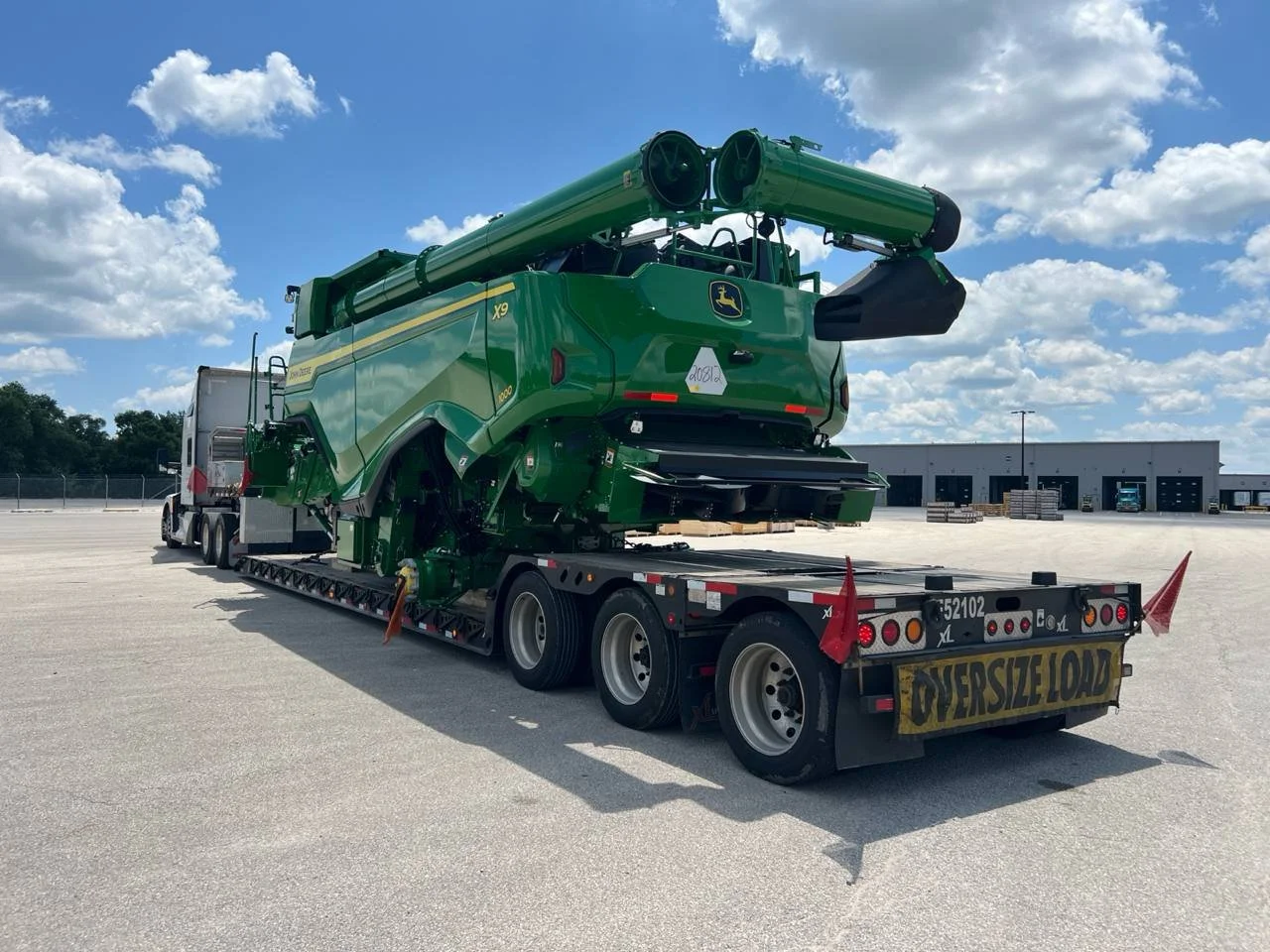 Large green John Deere combine harvester being transported on a flatbed truck in an open lot with a warehouse in the background.