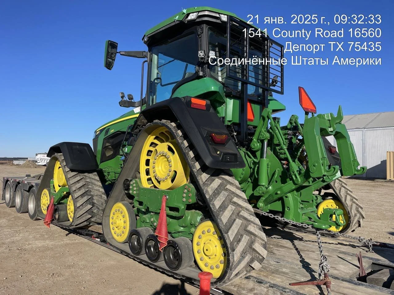 A green tractor with yellow wheels and black tracks, secured on a flatbed trailer, under a blue sky with an industrial building in the background.