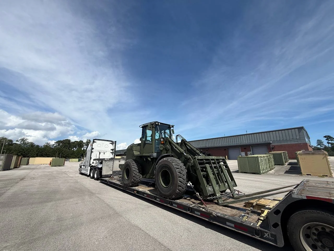 A large military vehicle being transported on a flatbed trailer in an industrial area with containers and a warehouse under a partly cloudy sky.