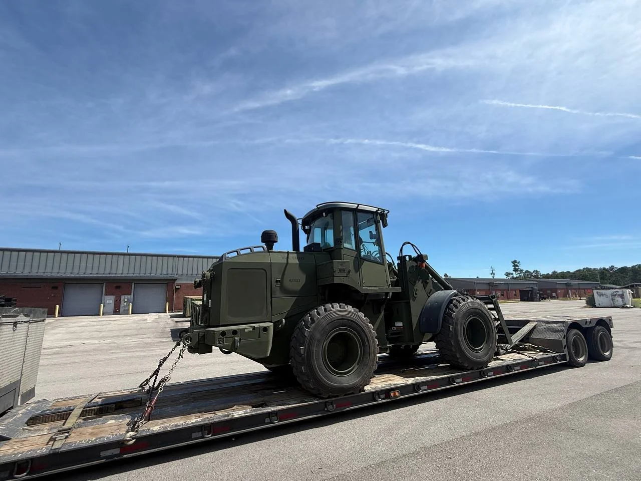 Military green tractor on a flatbed trailer, parked on an empty lot with warehouse buildings and a blue sky in the background.