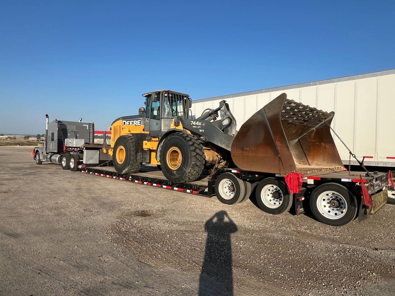 A large yellow and black John Deere wheel loader on a flatbed trailer, transported on a truck, with a white trailer and clear blue sky in the background.