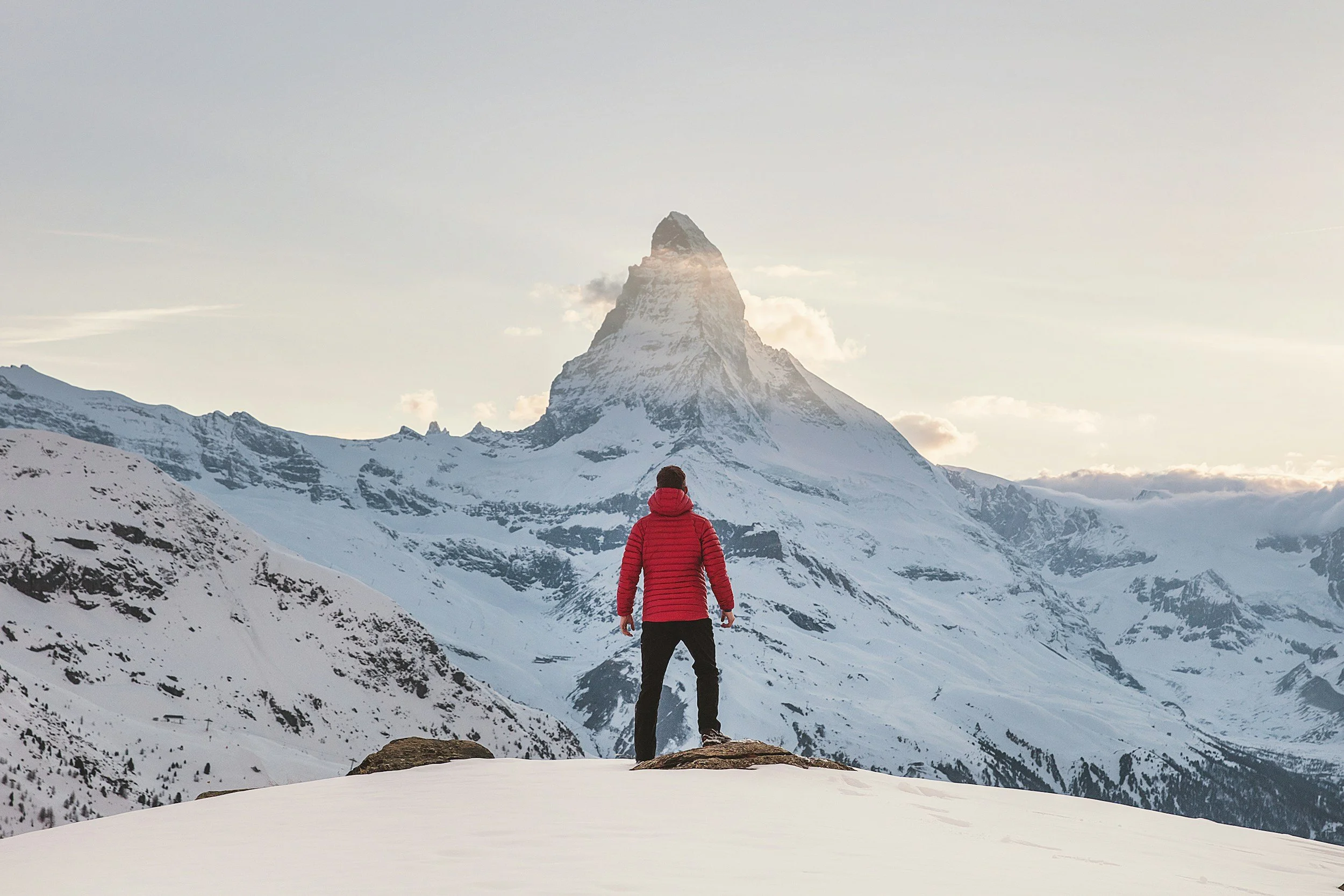 A person dressed in a red jacket and black pants standing on a snow-covered mountain, overlooking the snowy peaks and the Matterhorn mountain in the distance during sunset.