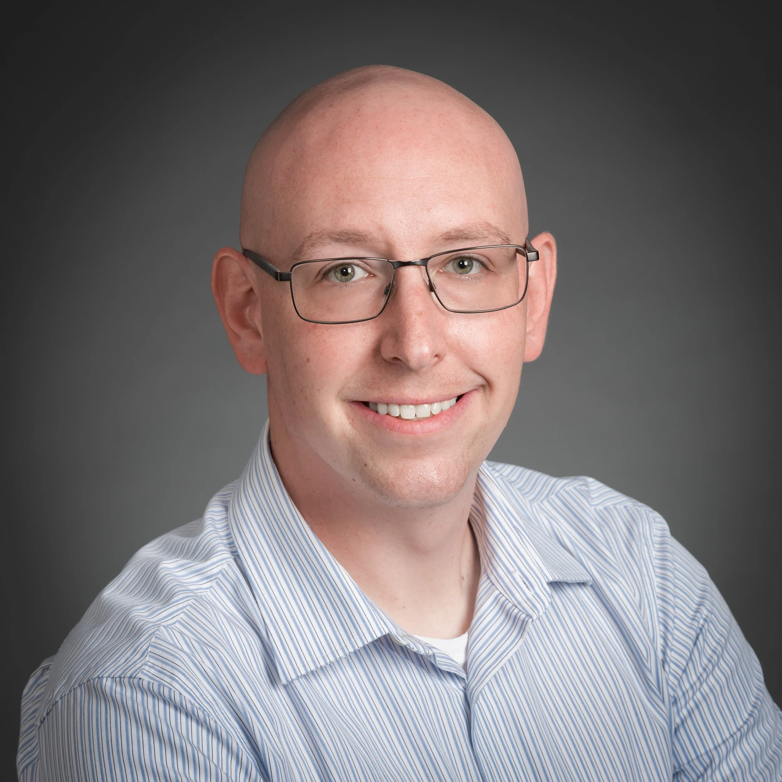 Professional headshot of a smiling man with glasses, bald head, wearing a striped collared shirt, against a gray background.