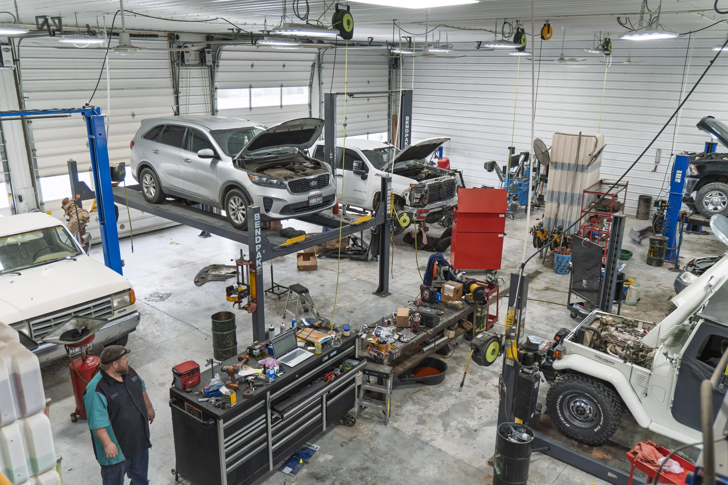 Automotive repair shop with multiple vehicles being serviced. One vehicle is on a lift with its hood open. The shop has tools, equipment, and workers working on cars.