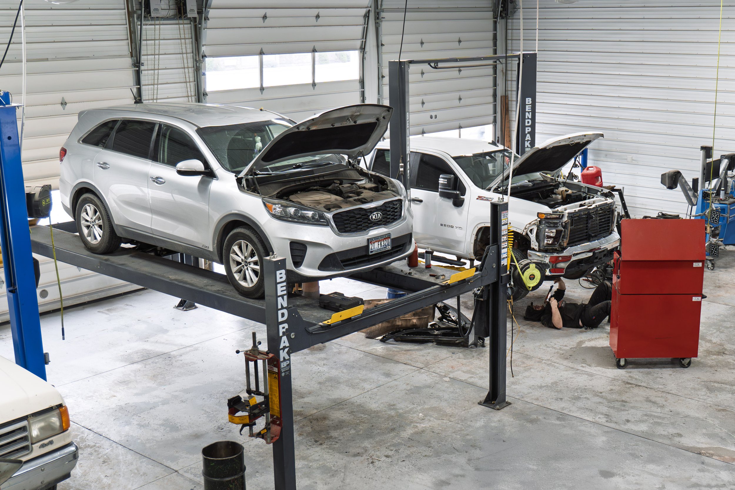 Automotive repair shop with two vehicles on lifts. One vehicle is a silver SUV with its hood open, and the other is a white pickup truck also with its hood open. A mechanic is working underneath the pickup truck while another person is working nearby on the floor.