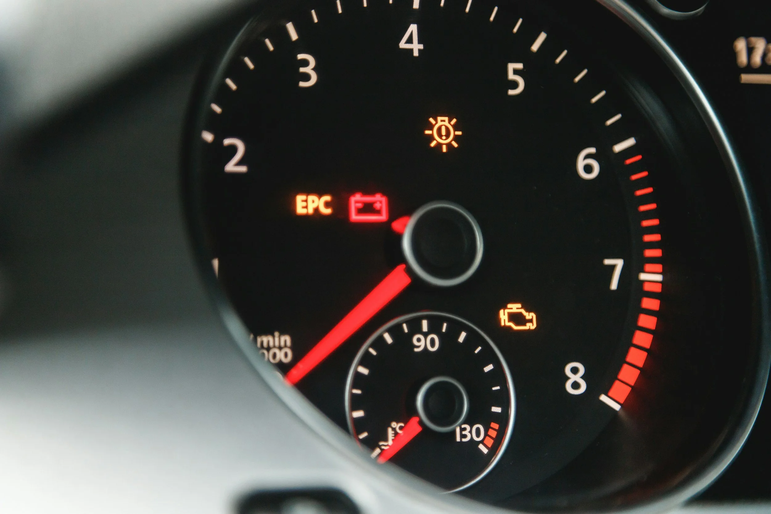 Close-up of a car dashboard showing warning lights including a brake warning light, a battery warning light labeled 'EPC,' and a check engine light with a yellow engine outline, with the tachometer needle pointing just above 1,000 RPM.