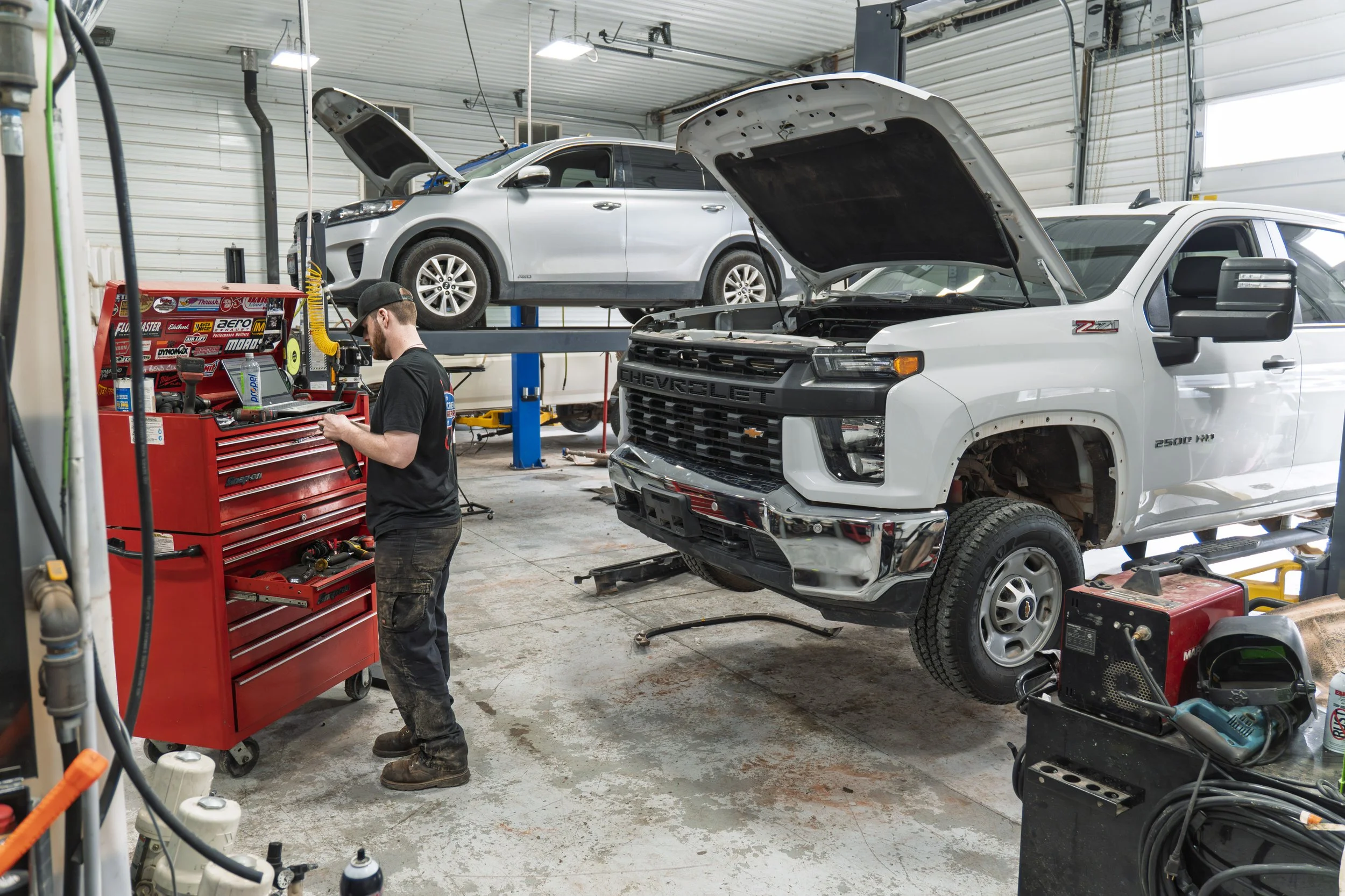 An auto repair shop with a mechanic working on a vehicle's engine. Two pickup trucks are elevated on hydraulic lifts, with both hoods open. The shop contains various tools, equipment, and supplies.