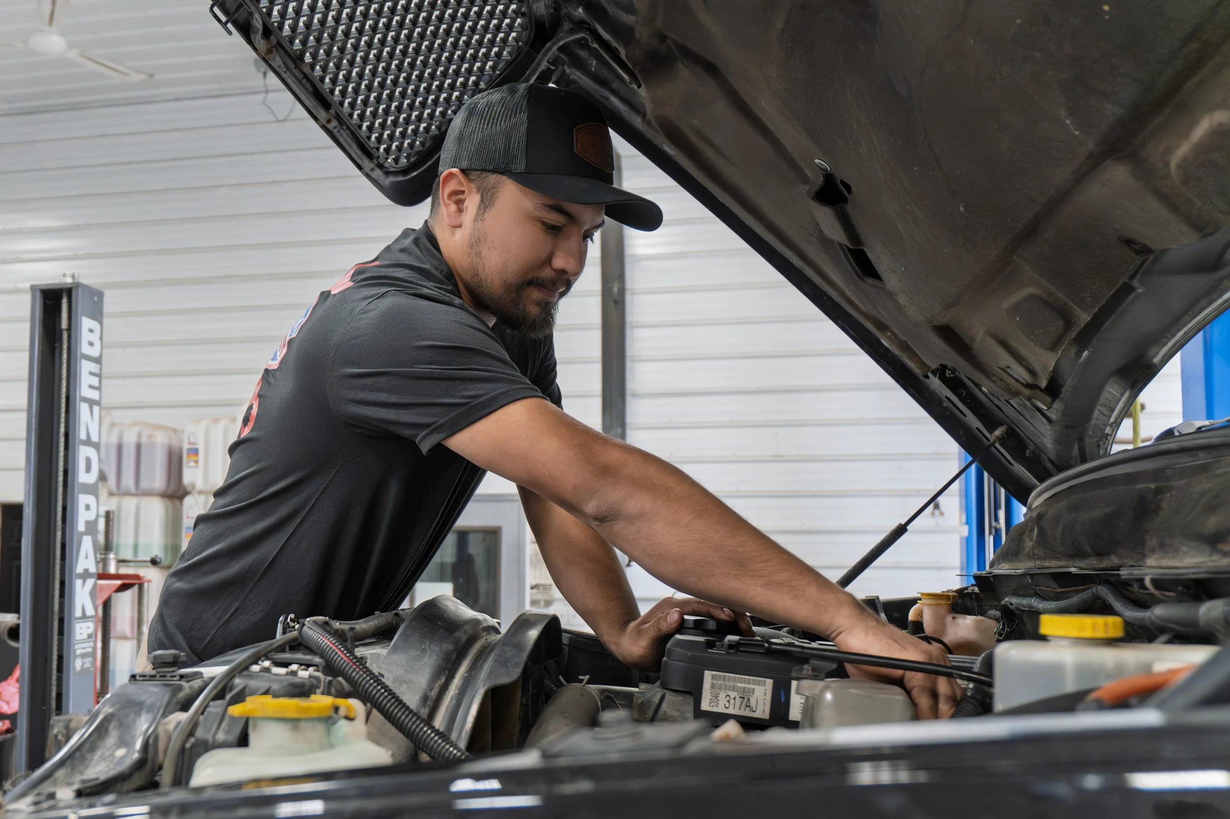 A man wearing a cap and black shirt inspected the engine of a vehicle inside a garage, with the hood open.