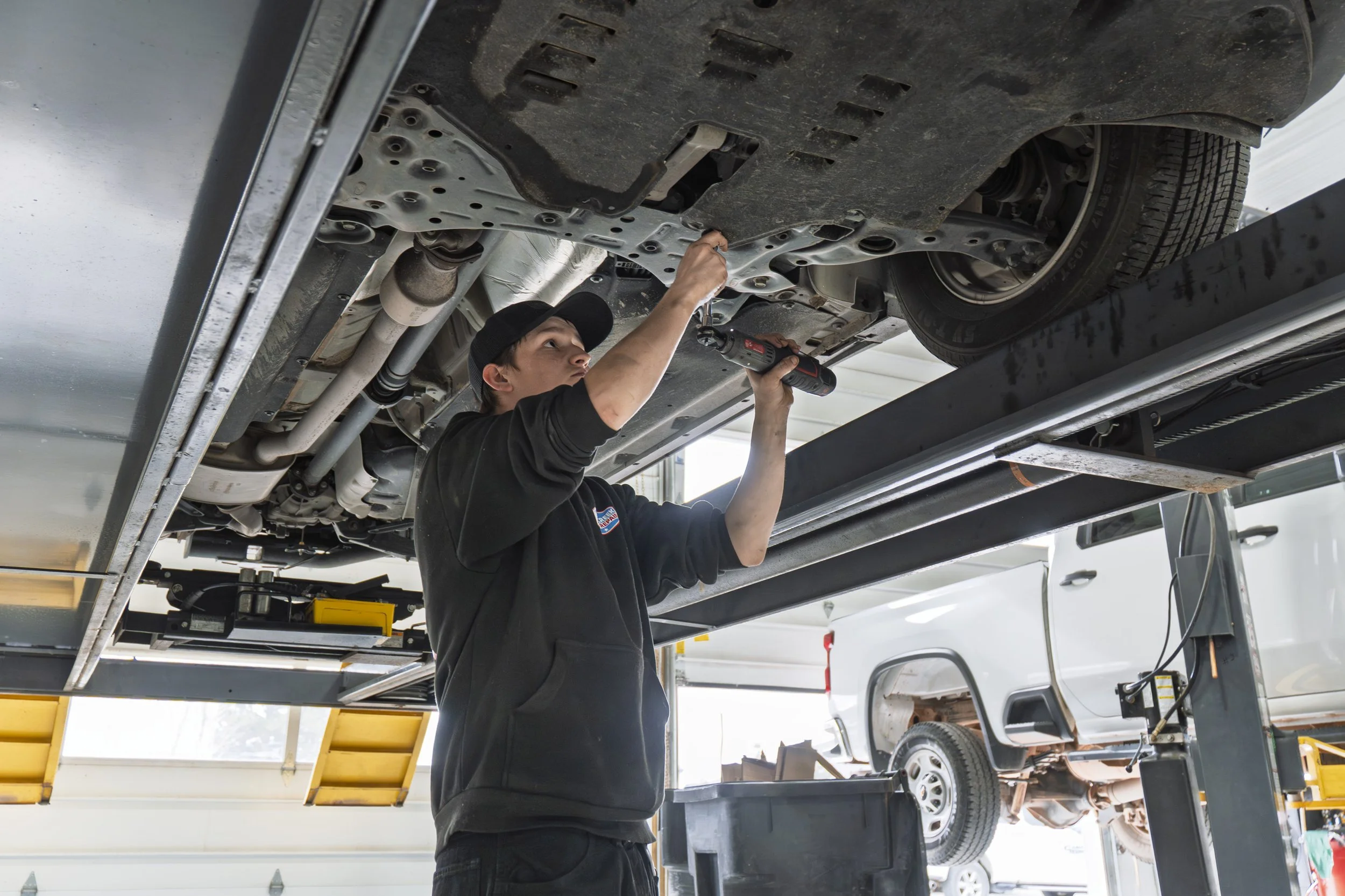 A person working underneath a vehicle on a hydraulic lift, using a tool to repair or inspect the car.