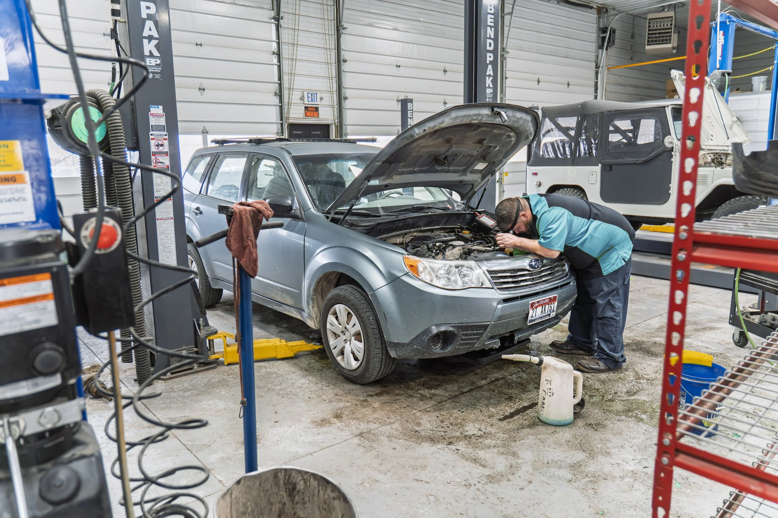 A mechanic inspecting the engine of a silver Subaru vehicle inside an auto repair shop. Another vehicle, a white off-road vehicle, is parked behind. The garage has a large roll-up door, tools, and equipment visible.