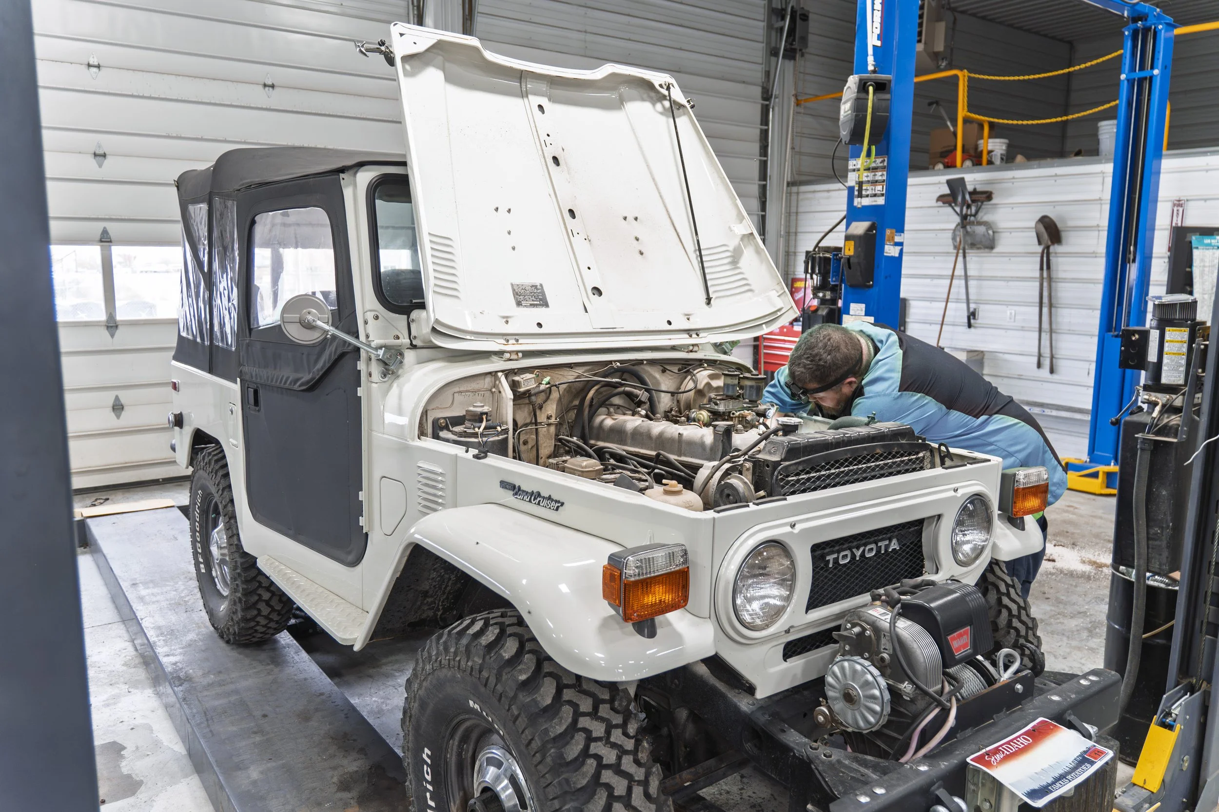 A person working on a white Toyota Land Cruiser with the hood open inside a garage, with tools and equipment around.