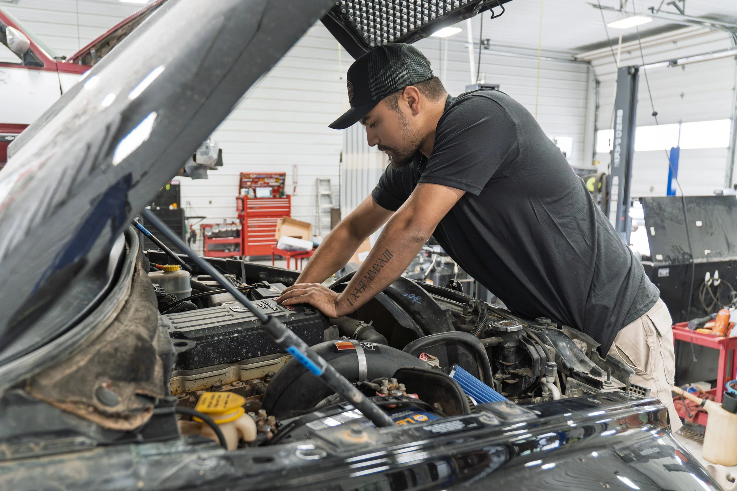 A man wearing a black baseball cap and black t-shirt working on a car engine inside a garage.