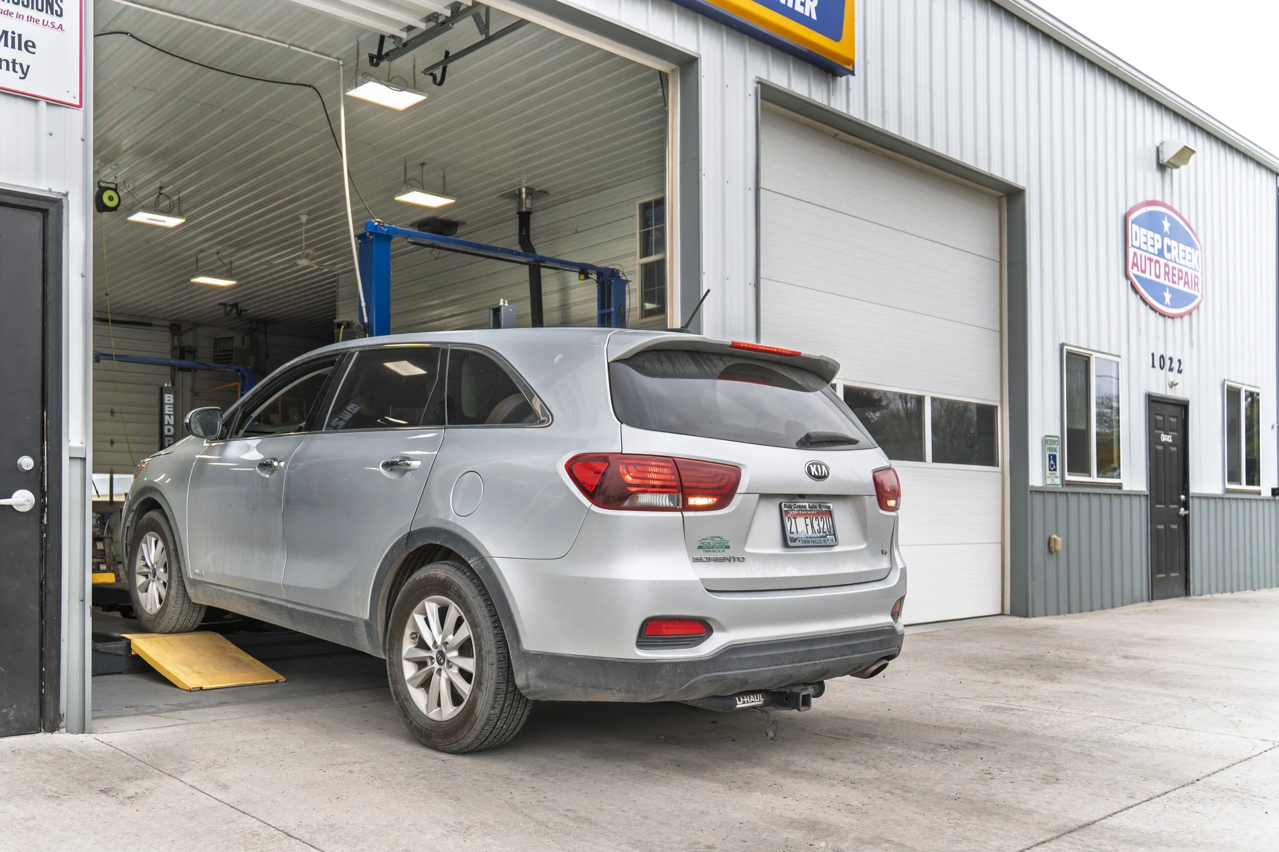 A silver Kia SUV on a vehicle lift inside an auto repair shop, with the rear wheels elevated on a yellow ramp. The shop has a large open garage door, and the exterior signage reads 'Deep Creek Auto Repair.' The building is white with vertical metal siding, and the shop's address is 1022.