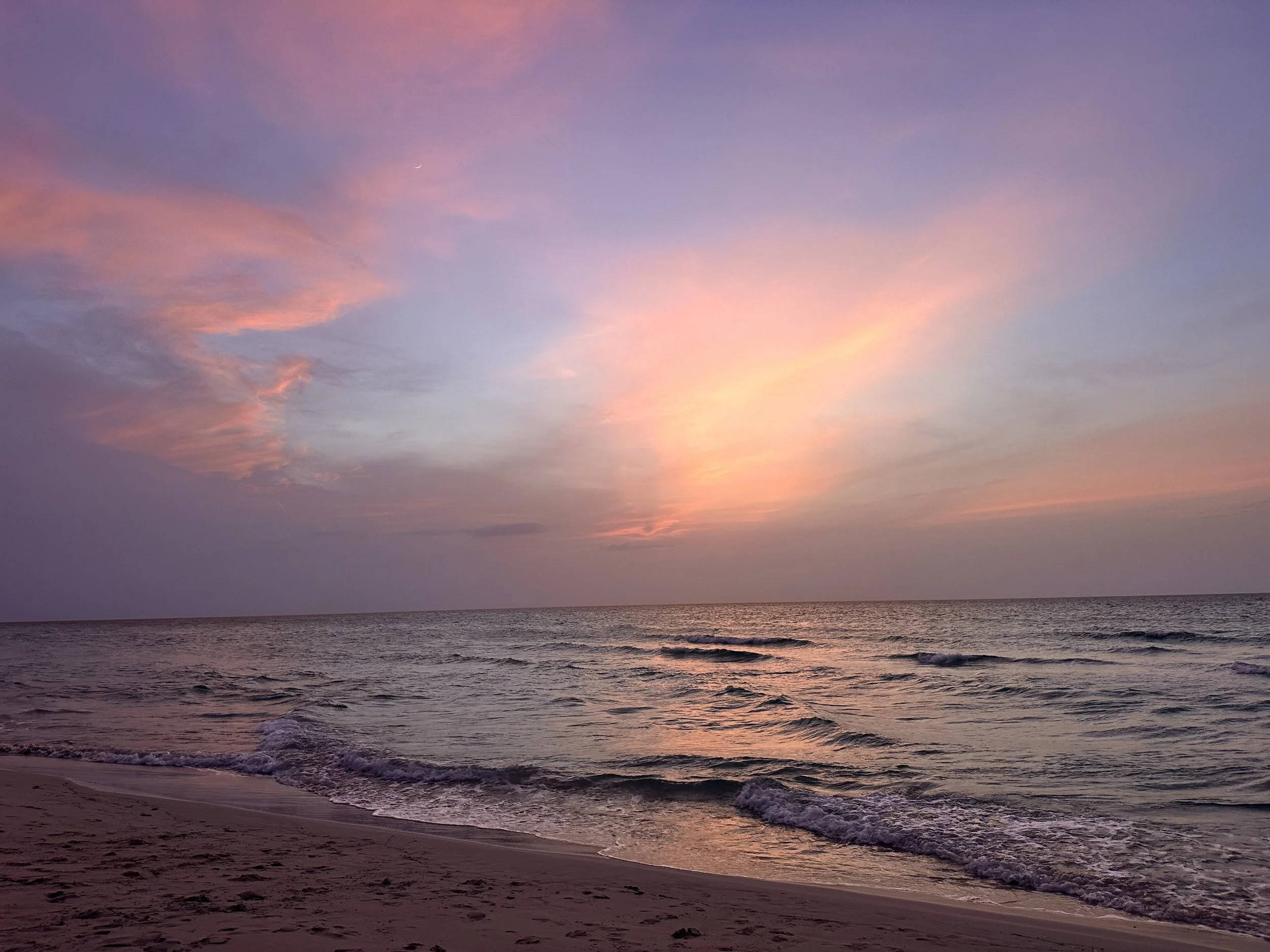 A peaceful beach at sunset with pink, purple, and blue clouds in the sky and gentle waves on the shore.