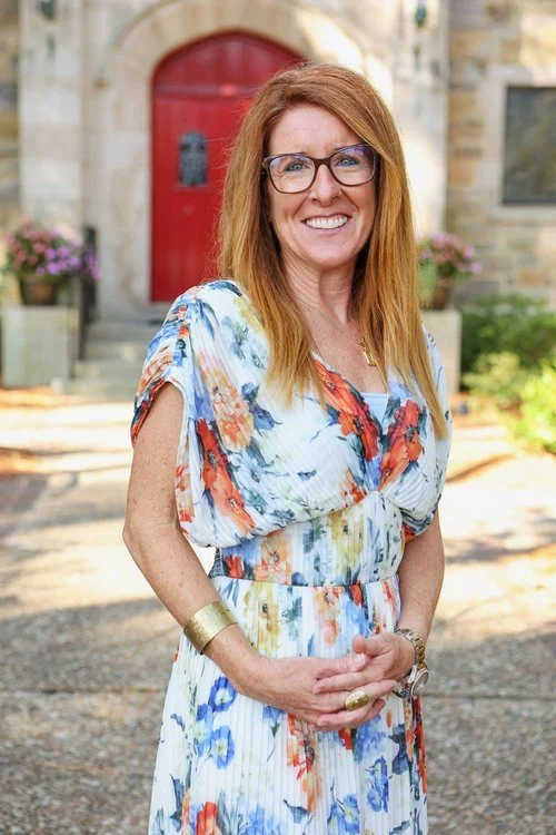 A woman with red hair, glasses, and a floral dress standing outdoors in front of a building with a red door and potted plants.