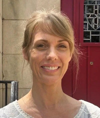 Close-up of a smiling woman with blonde hair in front of a stone wall and a red door.