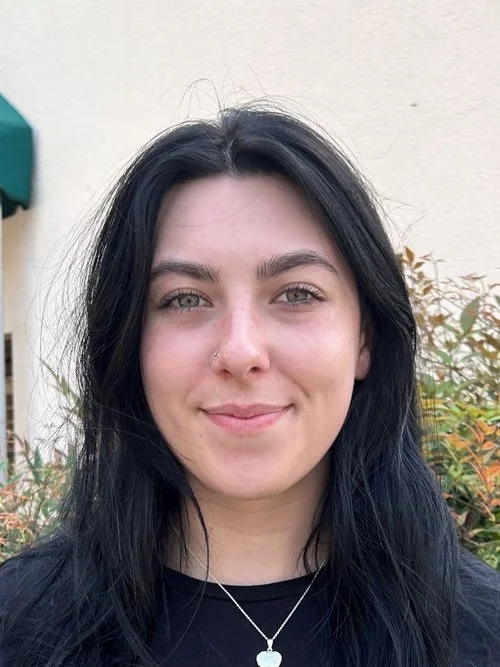 A young woman with long dark hair, light skin, and a nose piercing smiling outdoors against a background of plants and a beige wall.