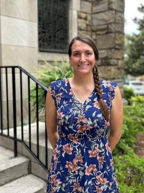 Woman with long braided hair wearing a blue floral dress standing outside in front of stone steps and greenery.