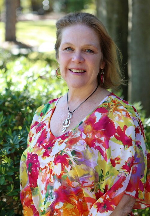 A smiling woman with light brown hair wearing a colorful floral blouse, standing outdoors with greenery and trees in the background.