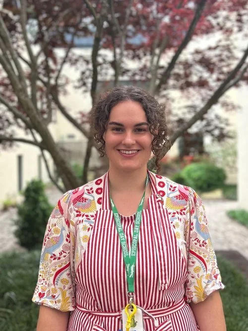 A woman smiling outdoors with pink and red blooming trees in the background, wearing a colorful floral and striped apron and a green lanyard.