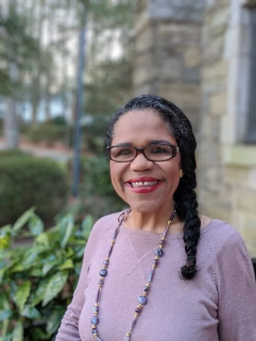 A woman with glasses and a braided hairstyle smiling outdoors with trees and a stone building in the background.