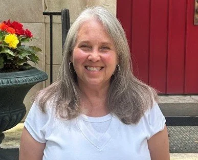 Smiling woman with gray hair wearing a white t-shirt, standing outside near a red door and a flower pot with red and yellow flowers.