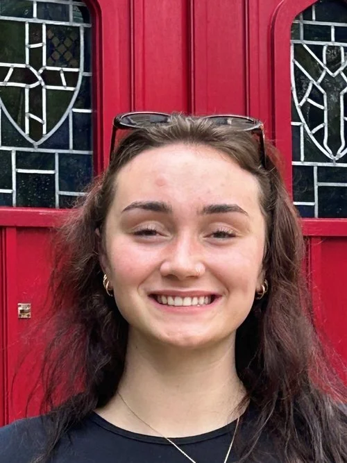A young woman with wavy brown hair, smiling, wearing glasses on her head, earrings, a necklace, and a black top, standing in front of a red door with decorative glass panels.