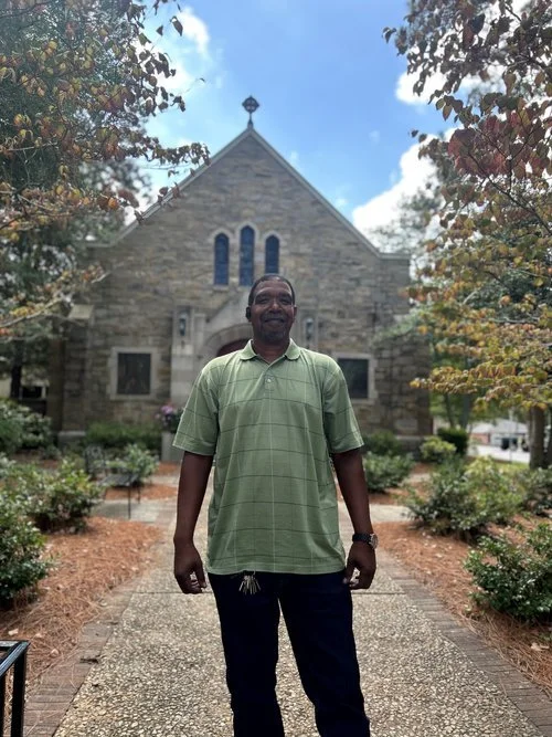 A smiling man in a green polo shirt standing in front of a stone church, surrounded by trees with autumn leaves.