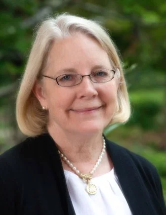A woman with blonde hair, glasses, wearing a black cardigan, white blouse, pearl necklace and earrings, standing outdoors with green trees in the background.