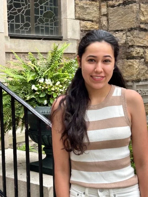 Young woman with long dark hair, wearing a sleeveless beige and white striped top, standing outdoors in front of a stone building and a potted plant with ferns and white flowers.