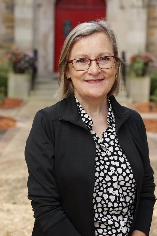 A smiling middle-aged woman with glasses and shoulder-length gray hair standing in front of a building with a red door and potted plants.