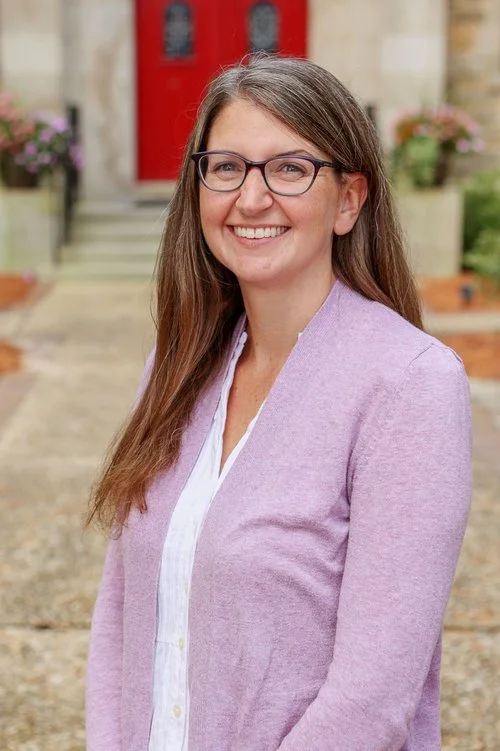 A woman with long brown hair and glasses smiling outdoors in front of a house with red door and potted plants, wearing a light purple cardigan over a white top.